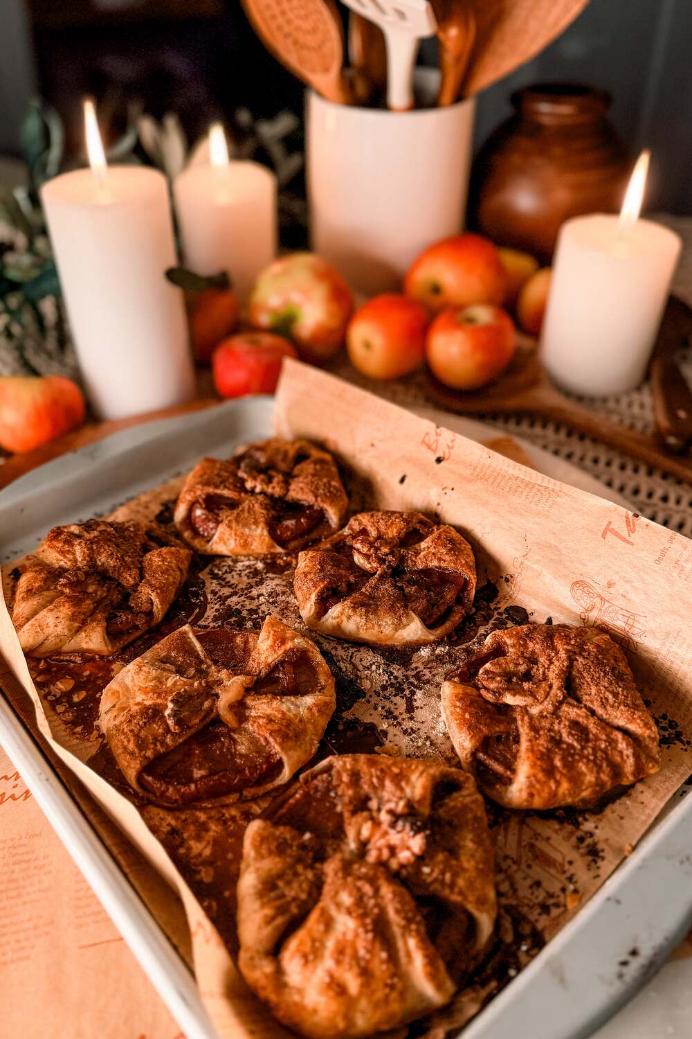 A sheet pan of golden baked apple puff pastry parcels on a table with lit candles and fresh red apples behind.
