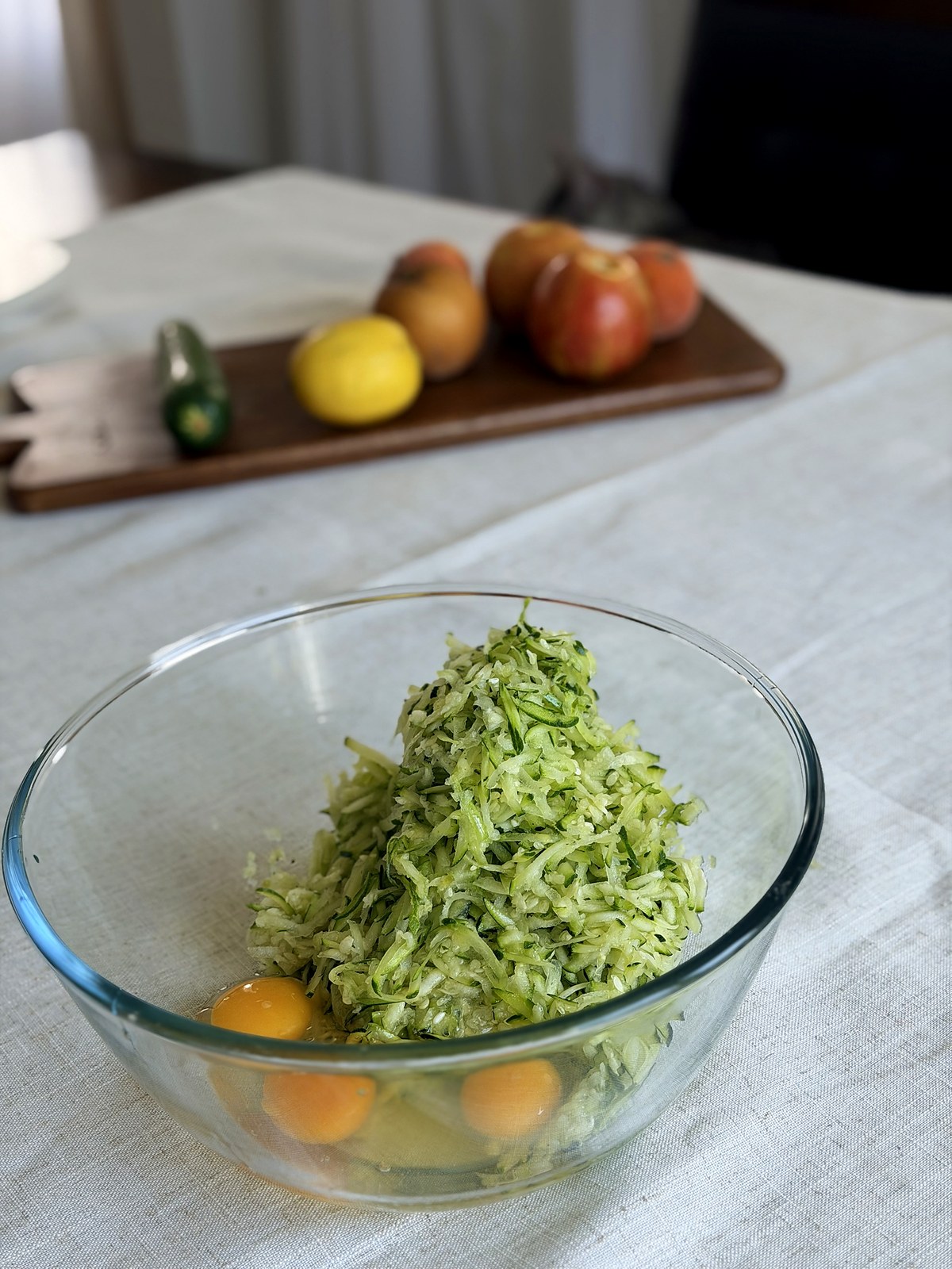 Grated zucchini piled in a large glass mixing bowl with three eggs at the bottom, on a linen tablecloth.