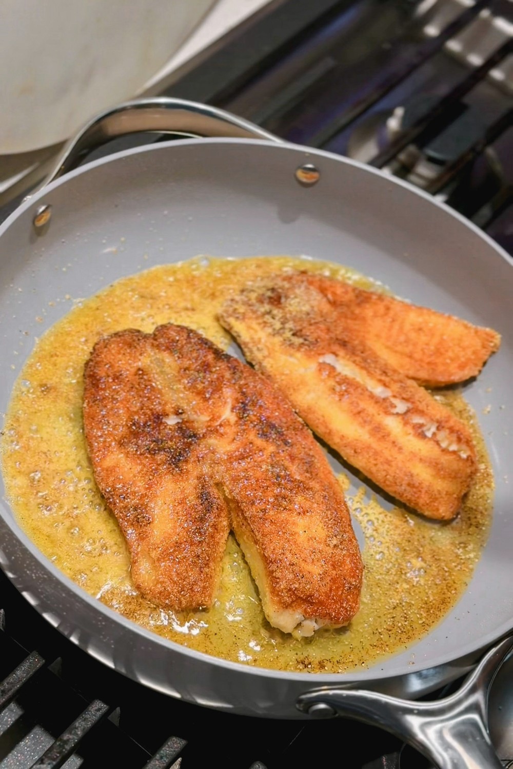 Two golden fish fillets cooking in butter in the Caraway ceramic sauté pan on a gas stove