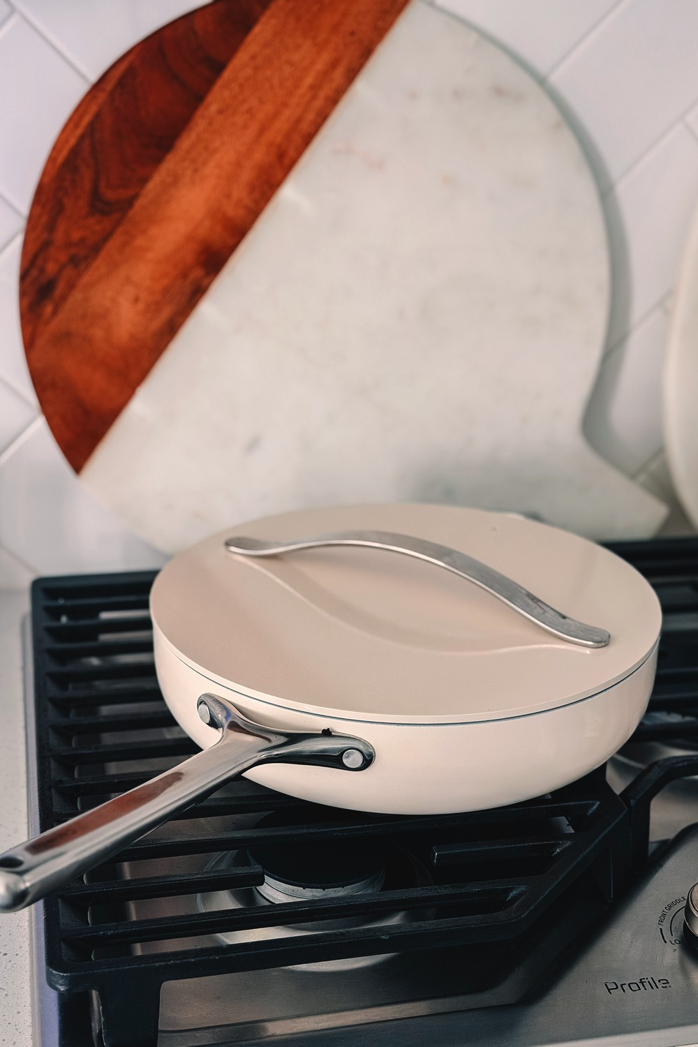 Caraway cream ceramic sauté pan with its lid on, sitting on a gas range in a white tiled kitchen