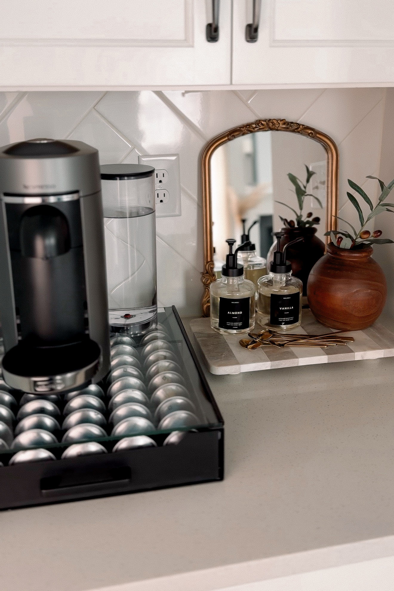 A small coffee bar styled on a kitchen counter with an espresso machine, a black tray of coffee pods, a gold arched mirror, glass almond and vanilla syrup bottles on a checkered marble tray, brass spoons and olive branches in a round wooden vase.
