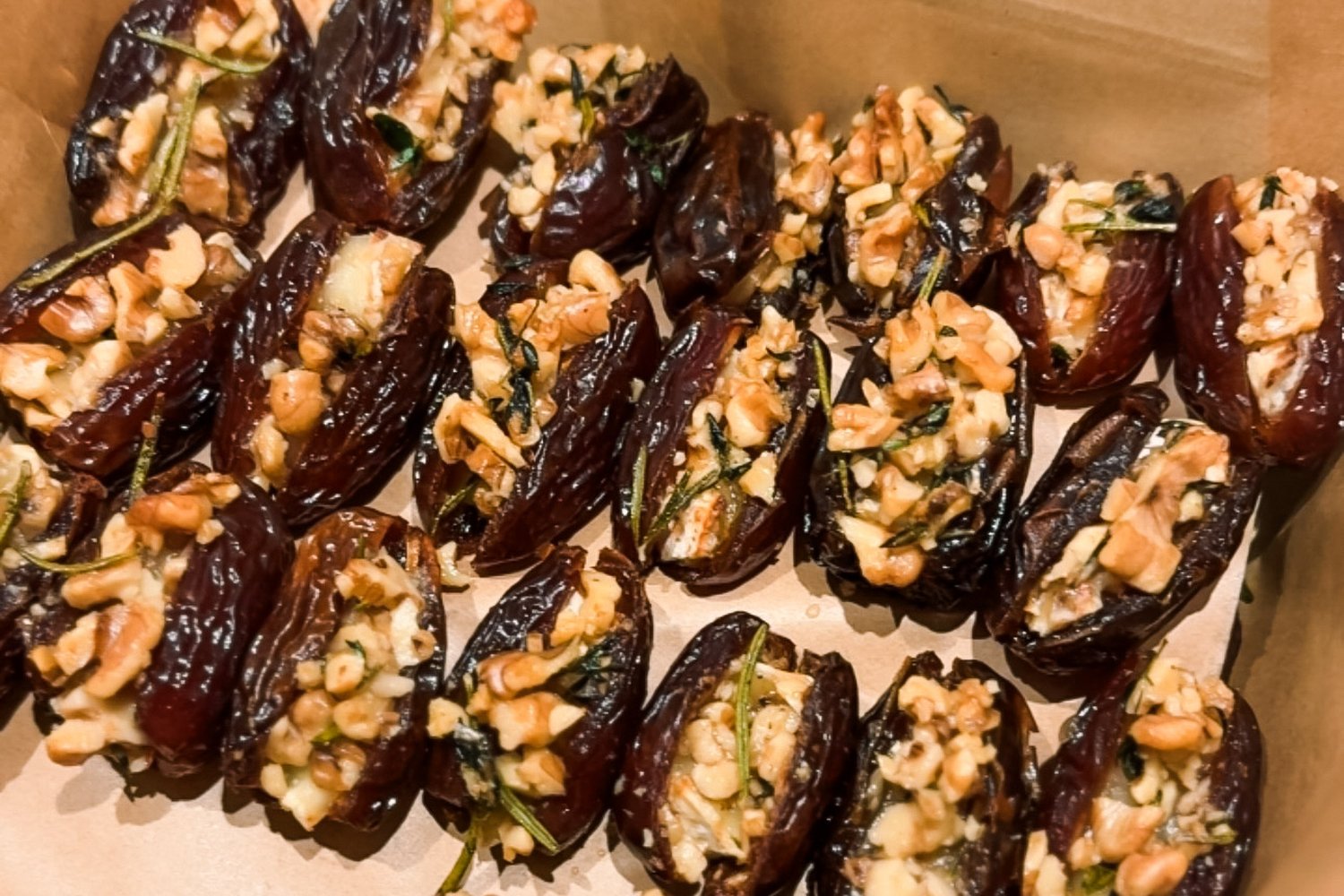 Overhead view of two dozen brie stuffed dates topped with chopped walnuts and rosemary, arranged on parchment paper in a baking pan before going into the oven.