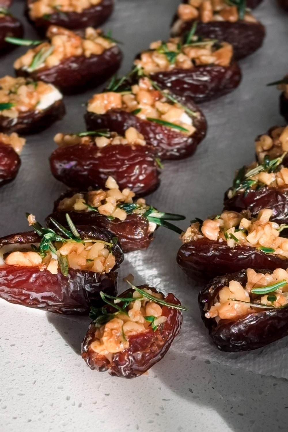Rows of baked brie stuffed dates topped with walnuts and rosemary on a dark tray, lit from the side.
