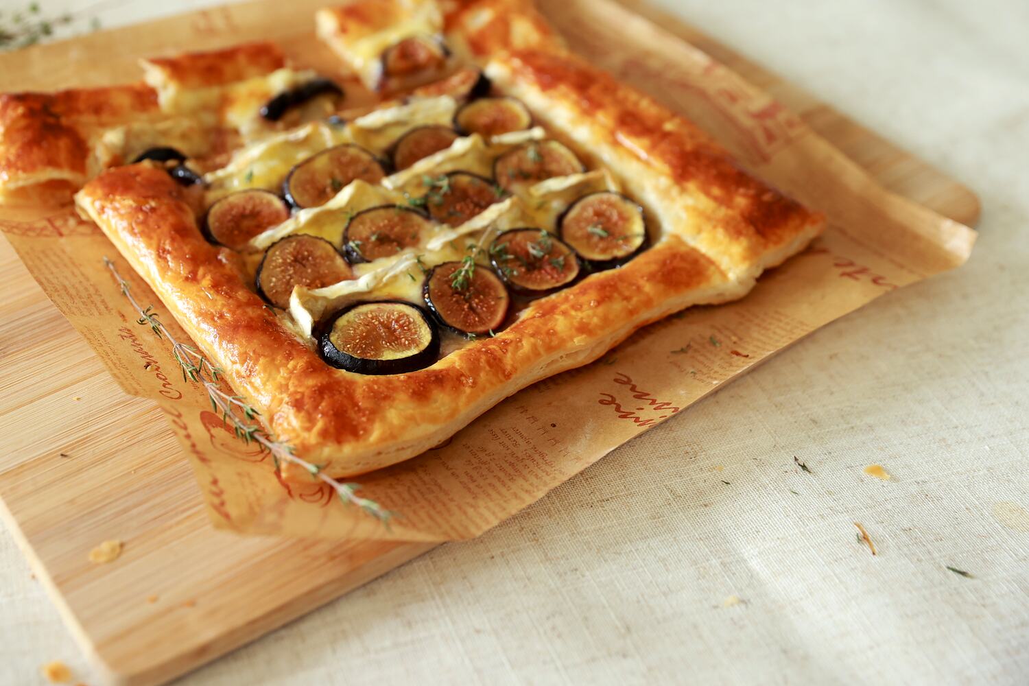 A baked fig and brie puff pastry tart on a wooden board with fresh thyme scattered around it.
