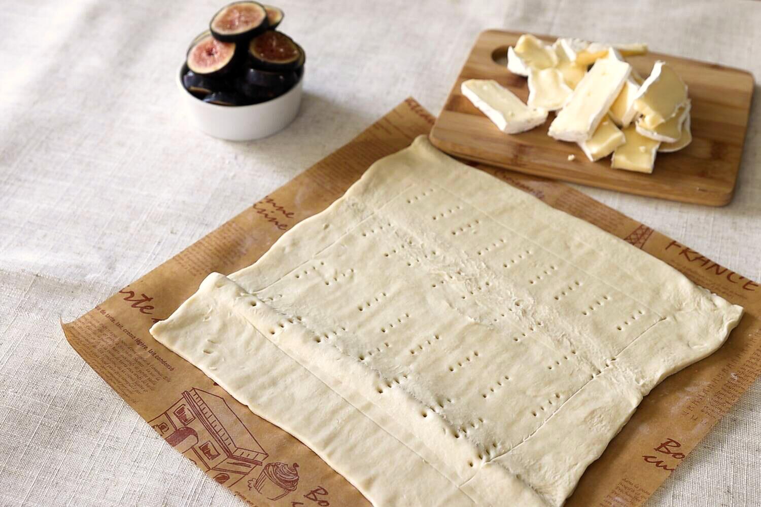Puff pastry docked with a fork on parchment, with sliced brie on a wooden board and fresh figs in a bowl behind it.