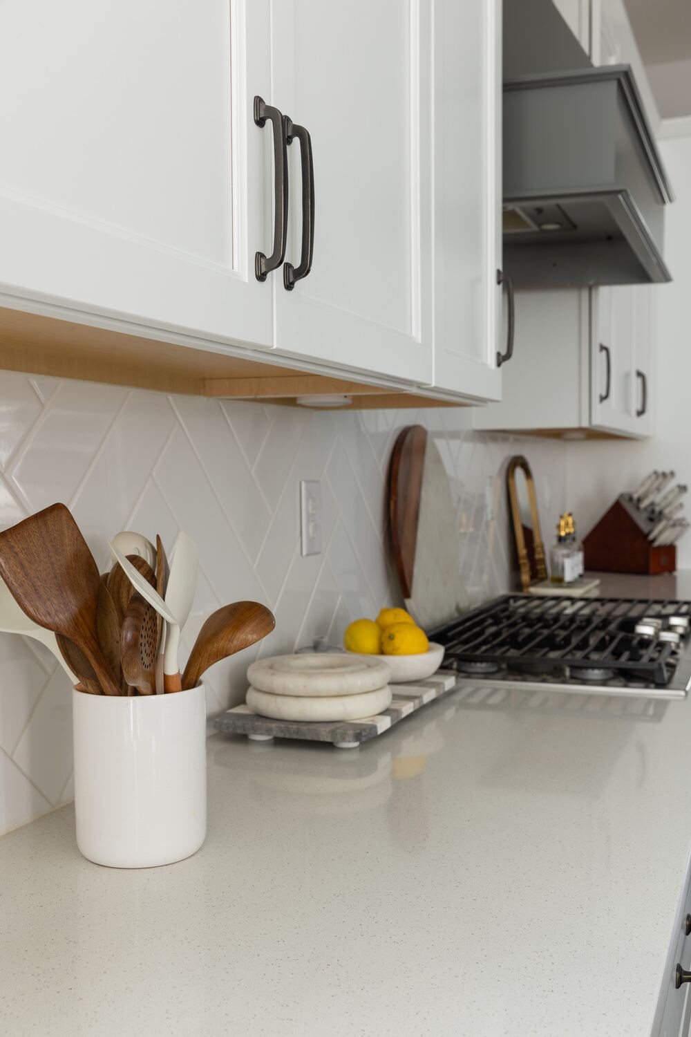 Wooden spoons in a white ceramic crock beside a bowl of lemons and stacked marble trivets on a white quartz counter.