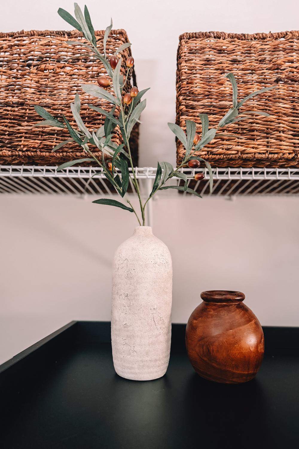 A cream ceramic vase holding a faux olive branch beside a small wooden pot on a black laundry counter.