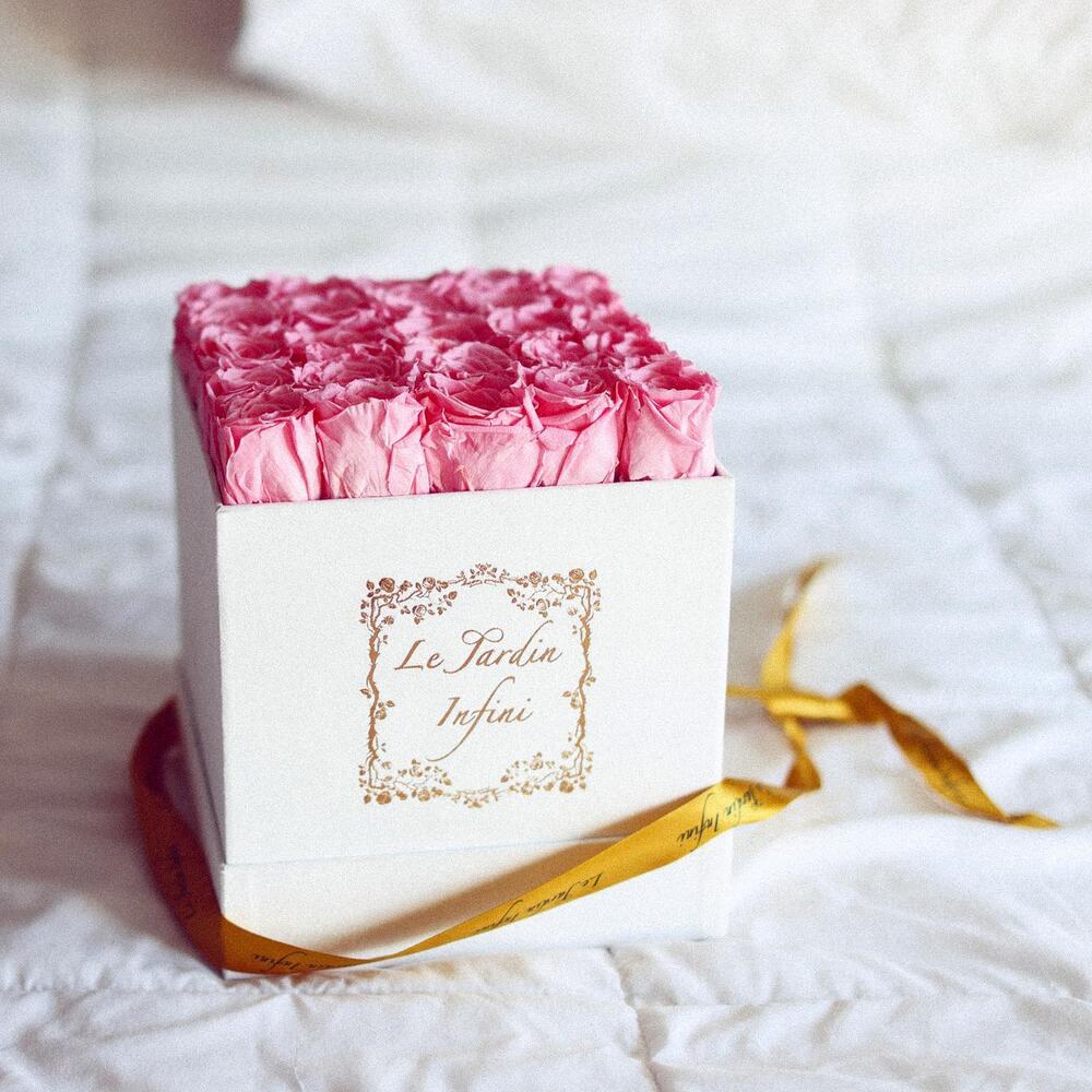 A white Le Jardin Infini box of preserved pink roses with a gold ribbon on white bedding.