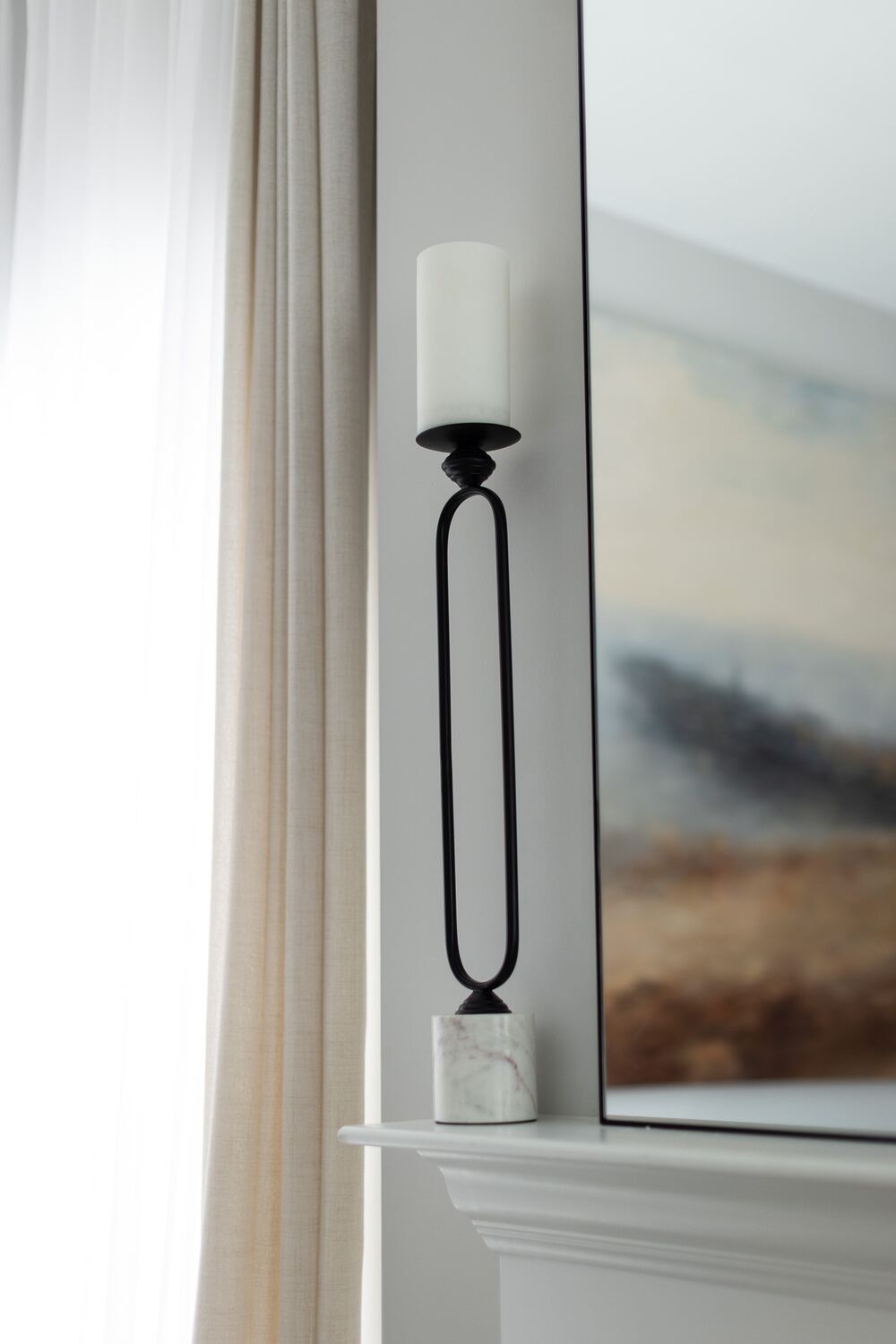 A tall black candlestick with a marble base on a white mantel, beside a linen curtain and the edge of a framed painting.