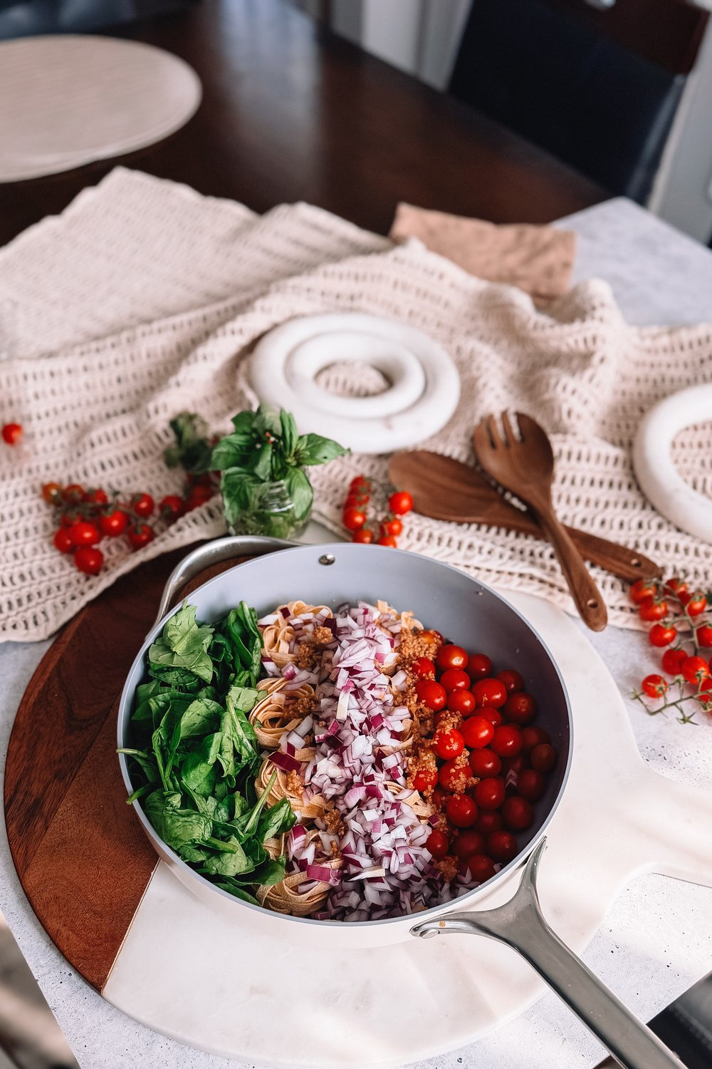 The filled skillet on a marble board on a kitchen table, with wooden serving spoons, vine cherry tomatoes and a small jar of basil around it.
