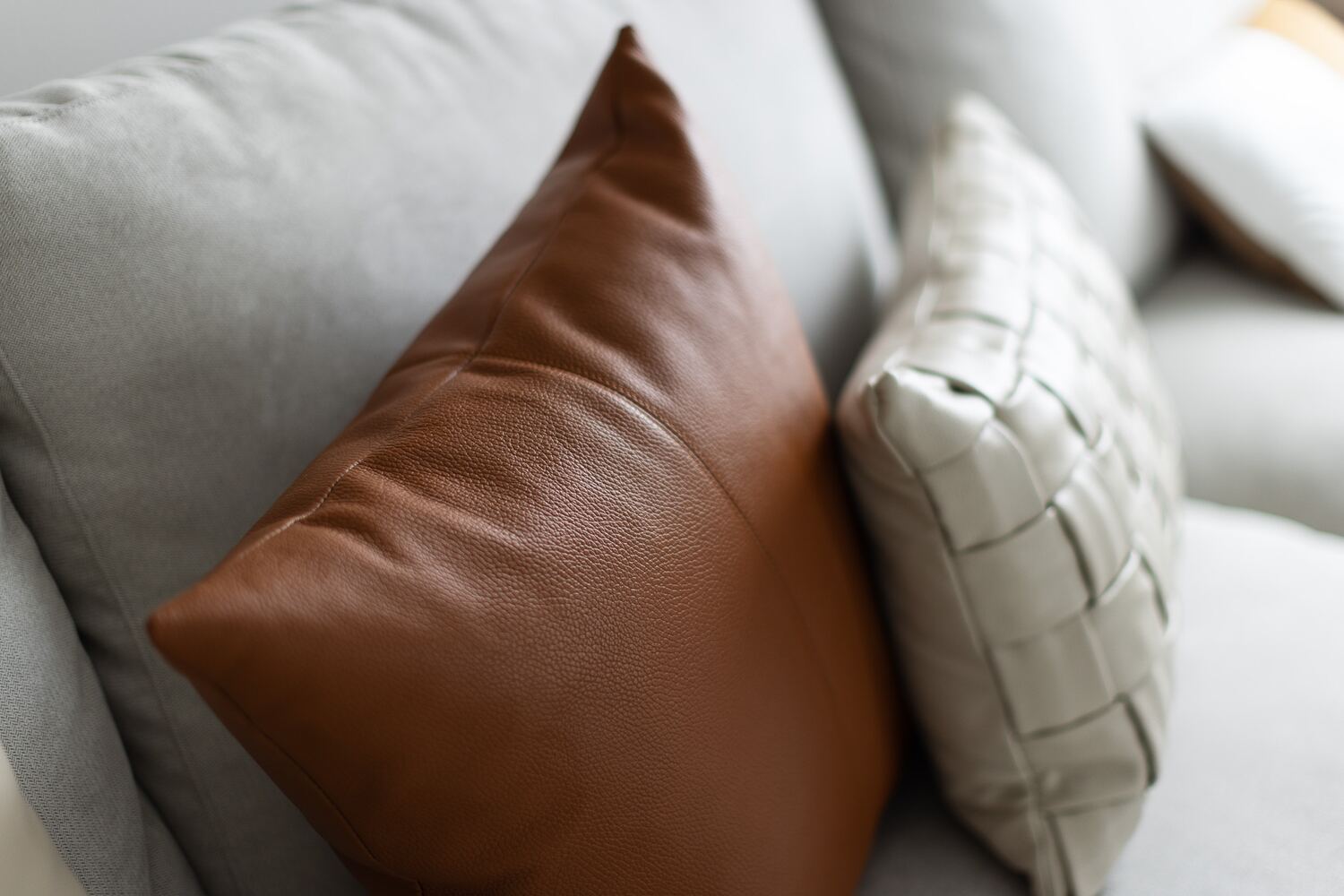 A tan leather pillow and a woven cream leather pillow resting on a pale grey linen sofa.