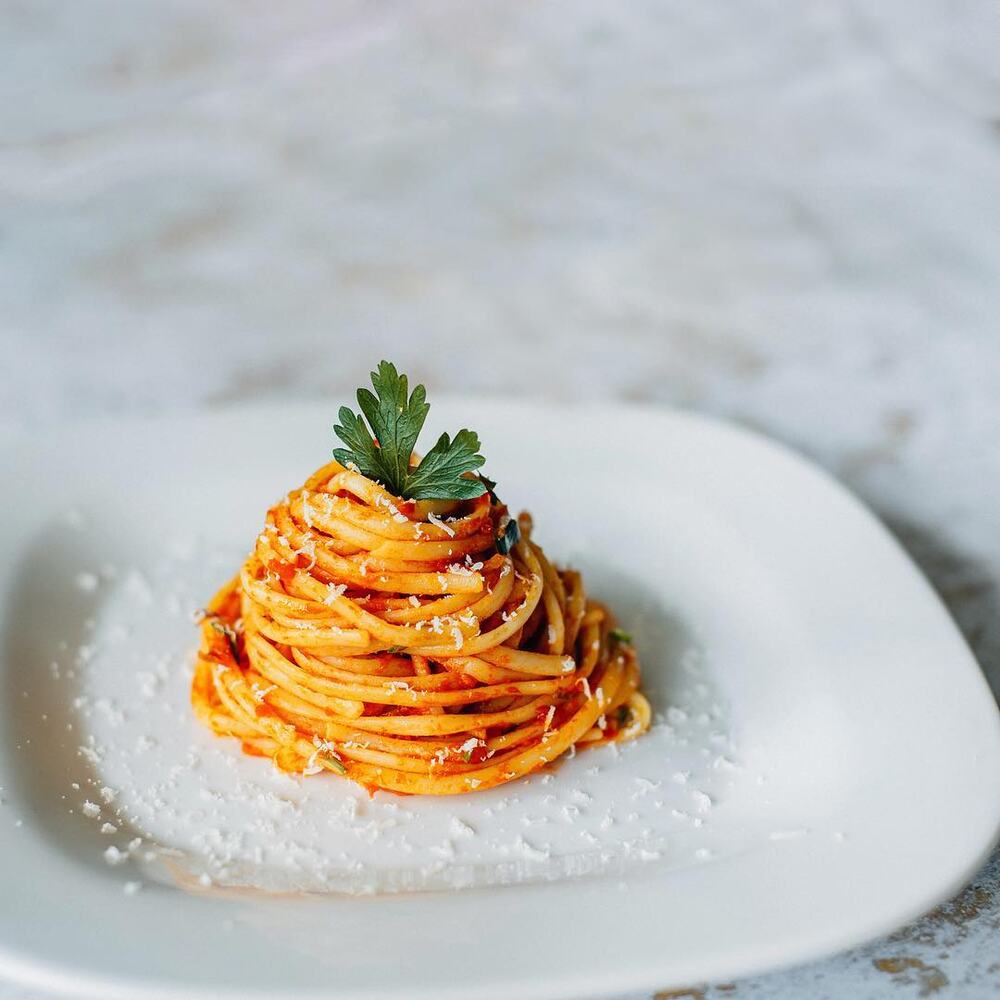A nest of bucatini in tomato sauce with parmesan and parsley on a white plate, shot on marble.