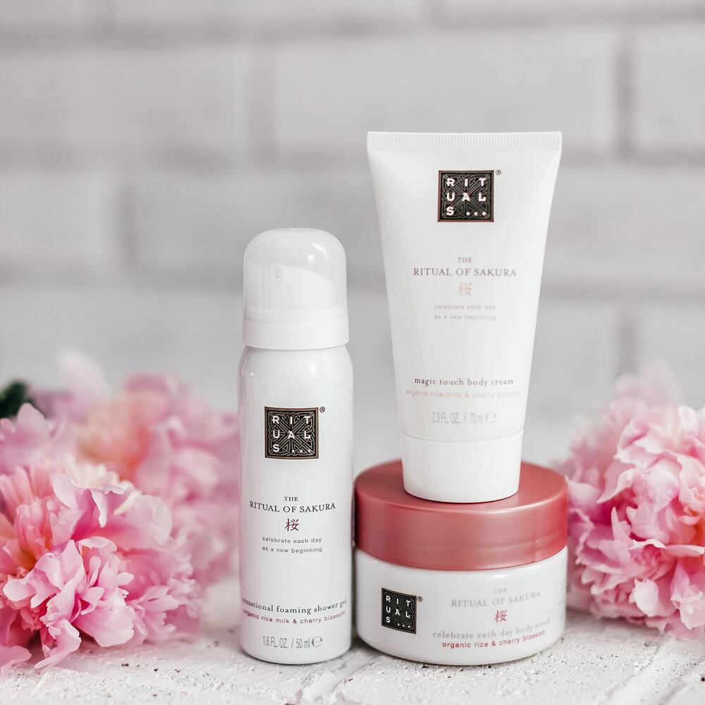 The Ritual of Sakura shower gel, body cream and body scrub arranged with pink peonies against a white brick wall.