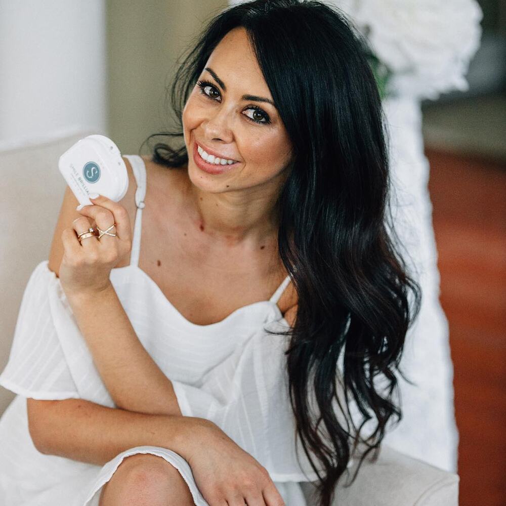 Olya Creative in white, smiling and holding a Smile Brilliant teeth whitening device.
