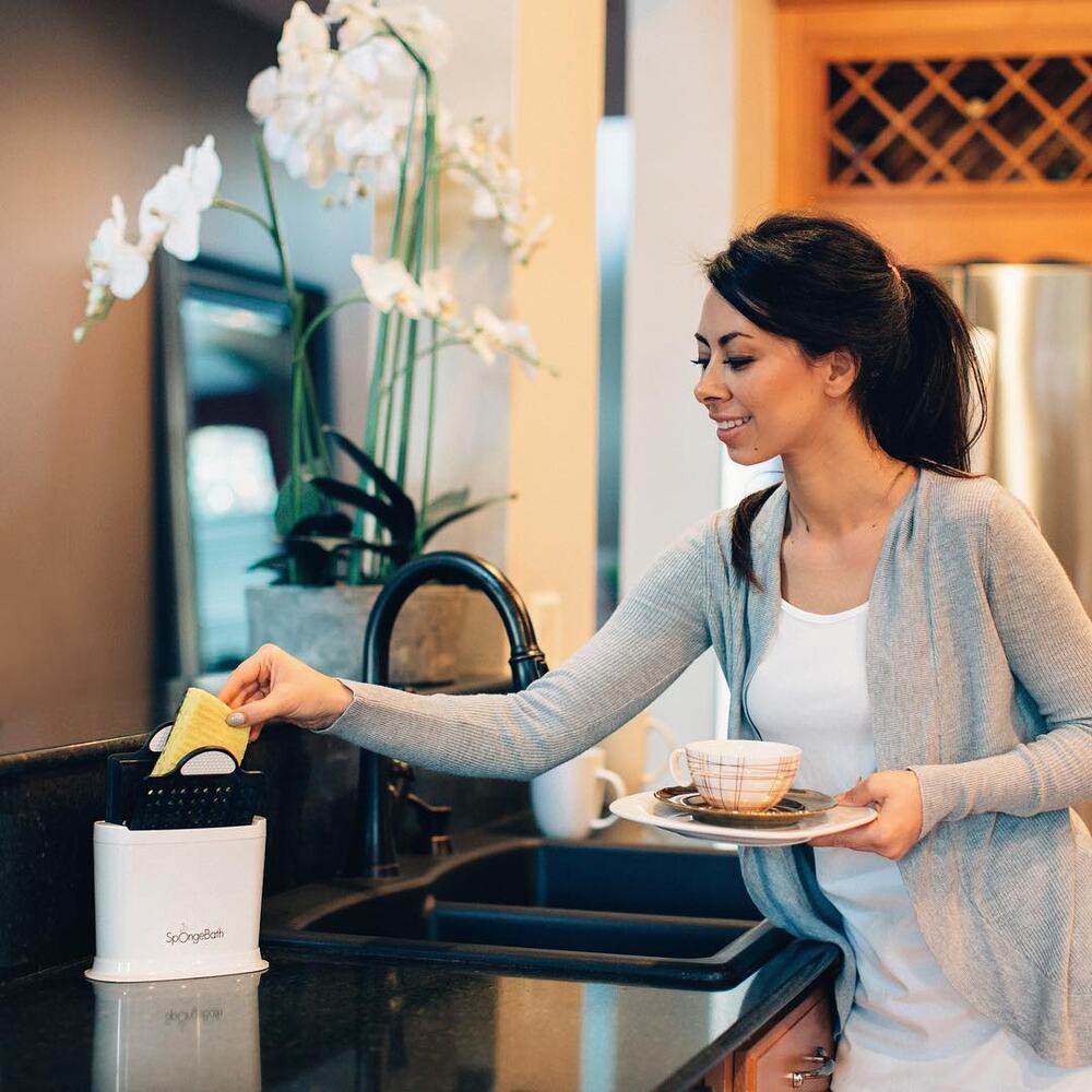 Olya Creative in her kitchen placing a sponge into a white SpongeBath caddy while holding a cup and saucer.