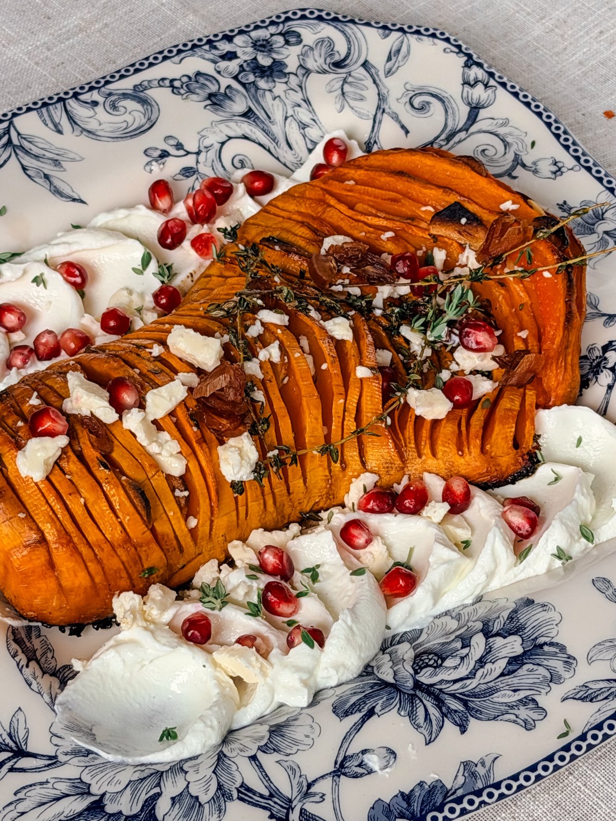 Hasselback butternut squash on a blue and white platter over yogurt, topped with feta, pomegranate seeds and thyme
