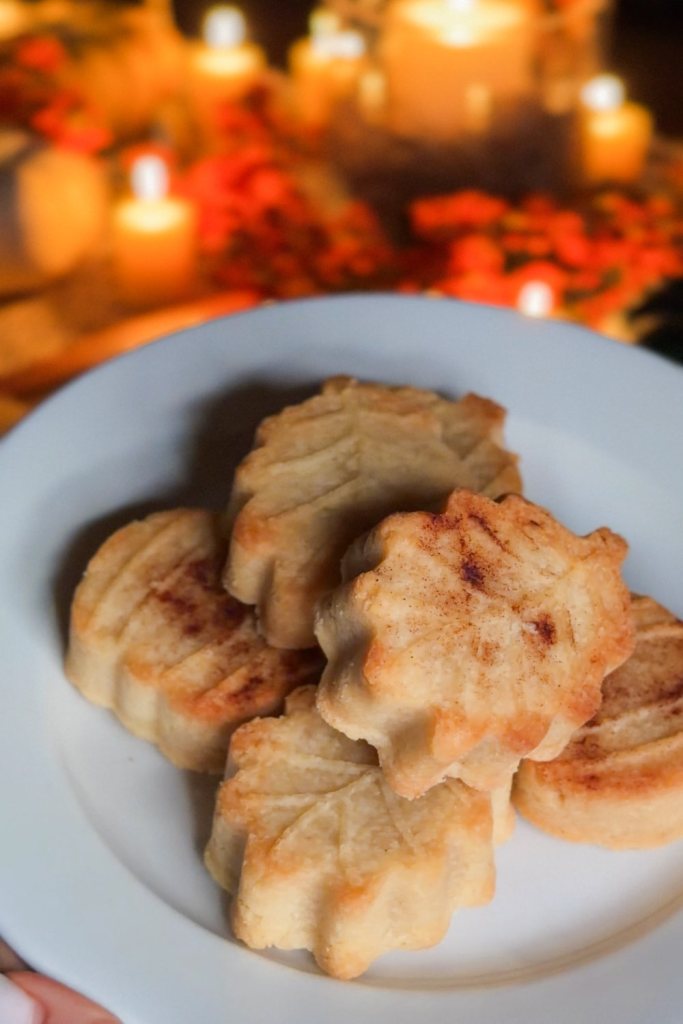 Baked leaf and pumpkin shortbread cookies on a white plate before dipping, with fall candles behind