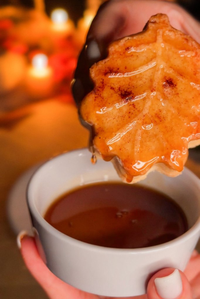 Oak leaf shortbread cookie being dipped into a white cup of warm maple syrup