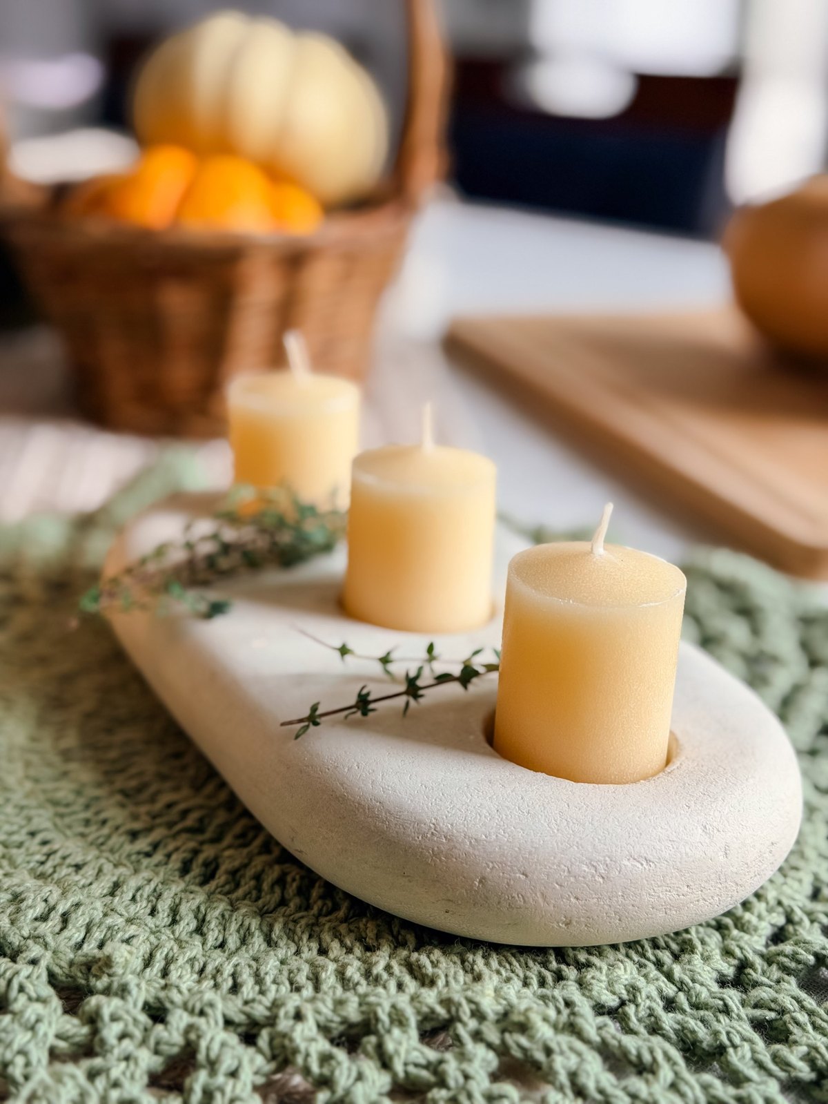 Three ivory pillar candles on a pale stone tray set on a green crocheted placemat, with small pumpkins and a basket behind