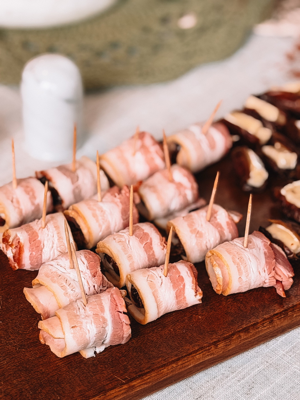 Dates wrapped in bacon and held with toothpicks, lined up on a wooden board before baking