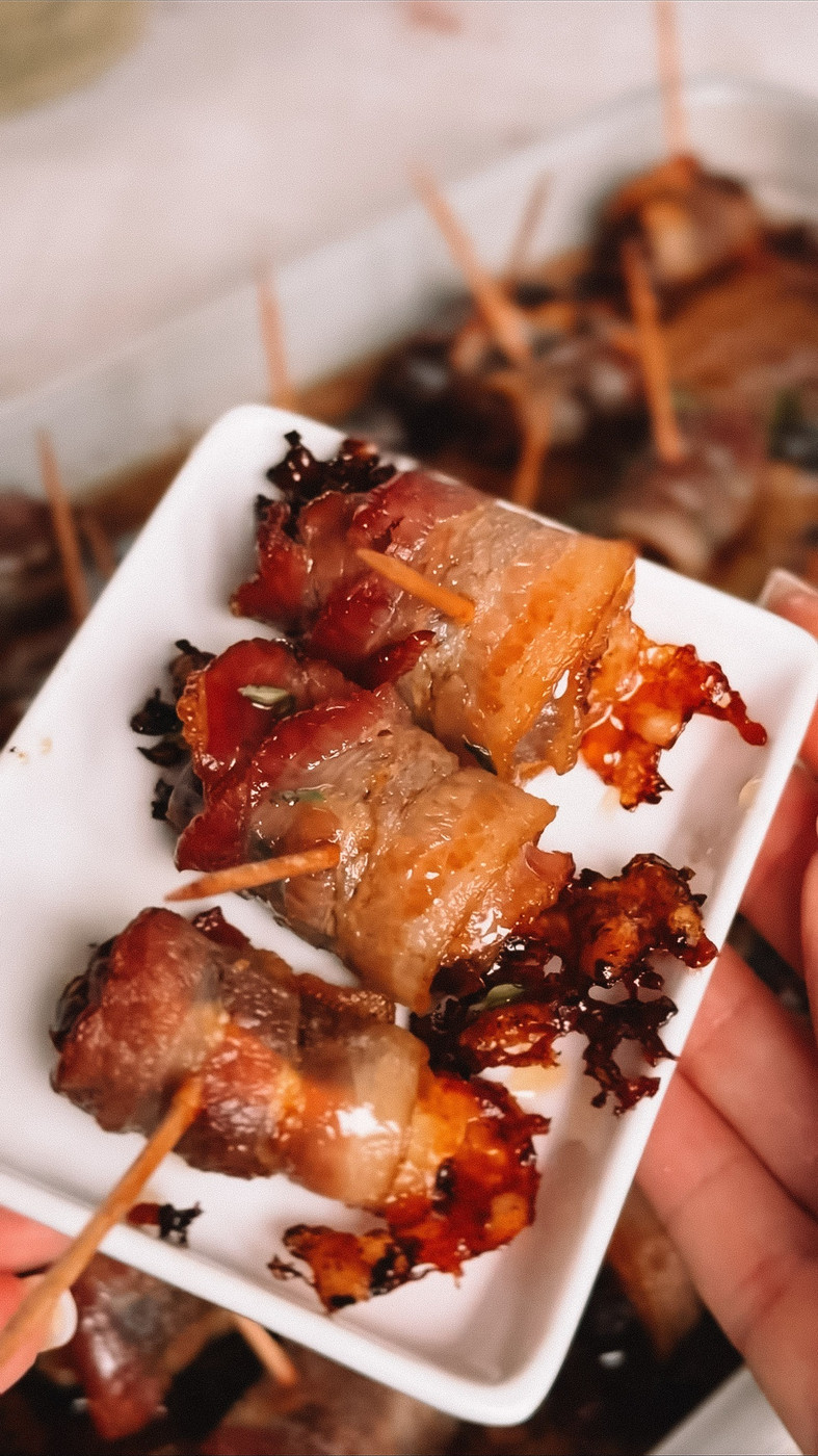 A hand holding a small white plate of baked bacon wrapped dates, with the baking dish behind