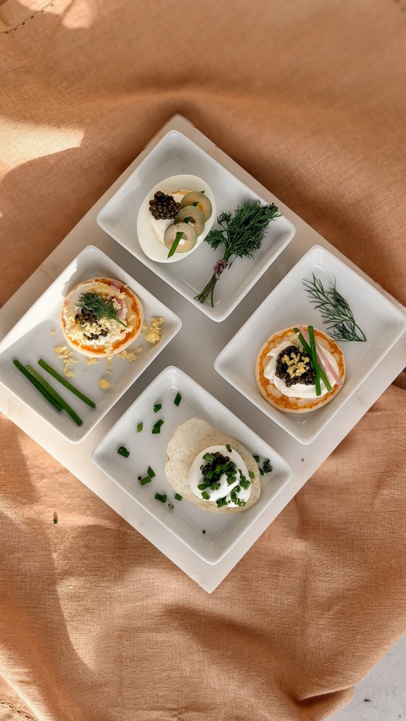 An overhead view of a four section white tray of caviar bites on tan linen in warm light.