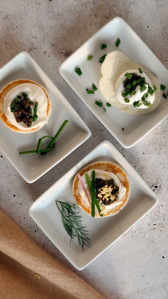 Three small white plates holding caviar on blini and caviar on a potato chip with cream and chives.