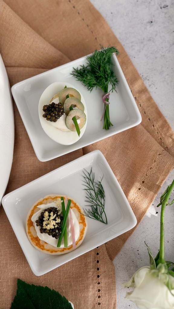 Two white plates, one with caviar on a halved egg and one with caviar on a blini with cream and chives, styled with dill.