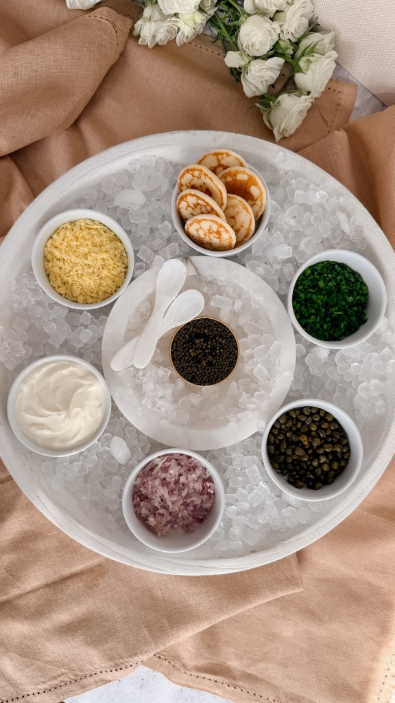 A round board of crushed ice with a tin of caviar and two mother of pearl spoons in the center, surrounded by small bowls of sieved egg yolk, blini, chives, capers, red onion and cream.