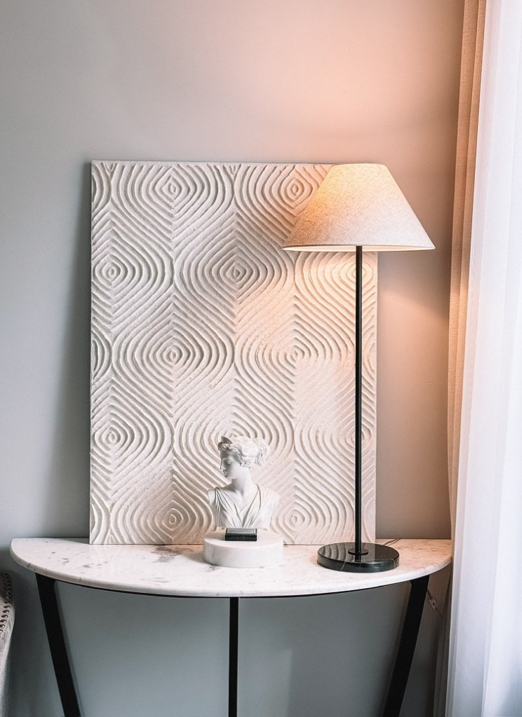 A half moon marble console table with black metal legs, a lit black lamp with a cream linen shade, a white bust on a round marble riser, and a carved geometric panel leaning against the wall.