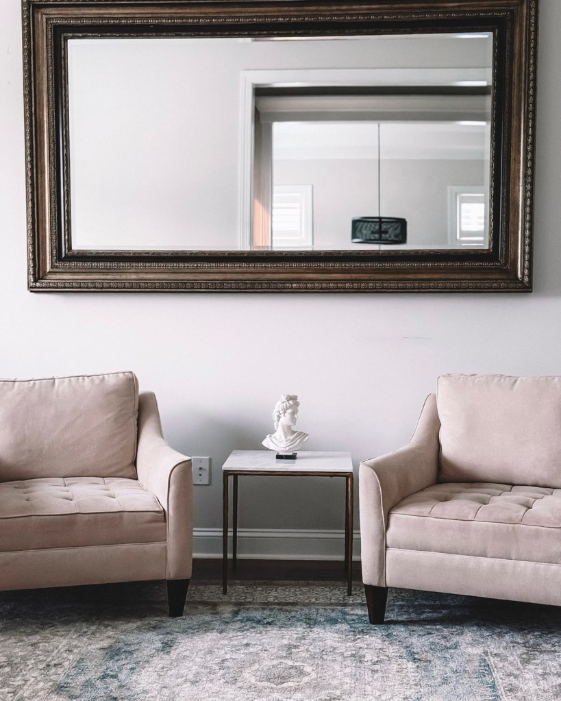 A wide rectangular mirror hung horizontally above two blush tufted armchairs, with a small marble side table and a white bust between them.