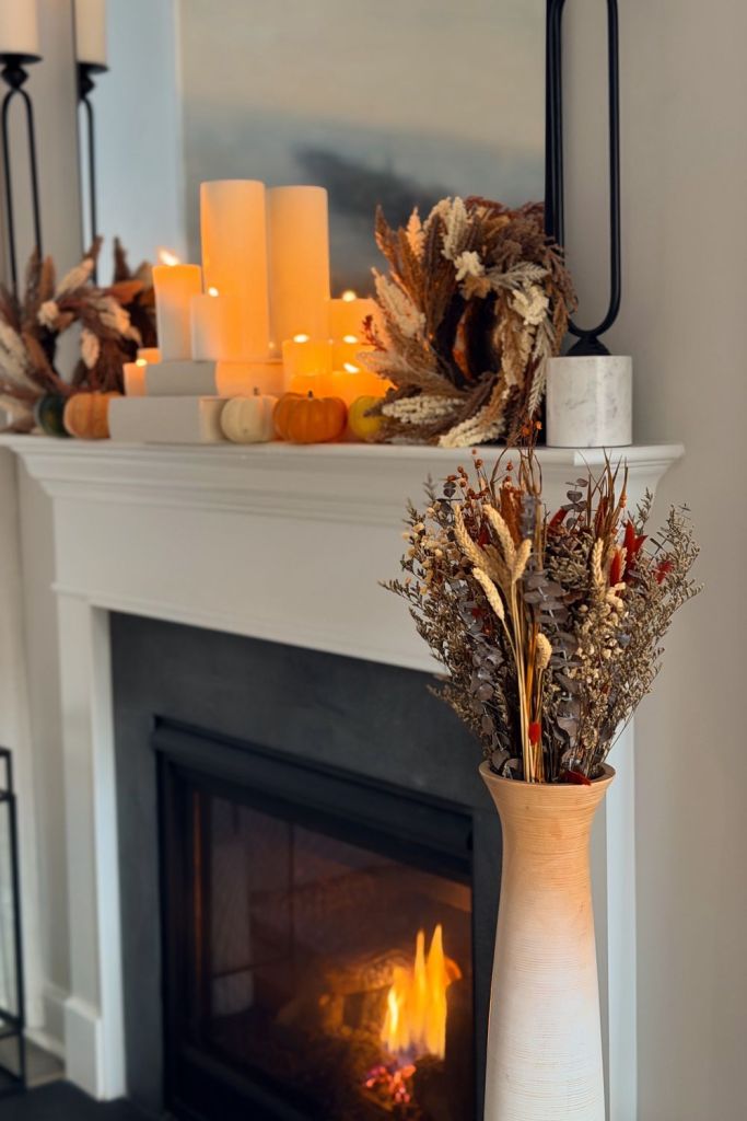 Dried fall stems in a tall cream vase beside a lit fireplace