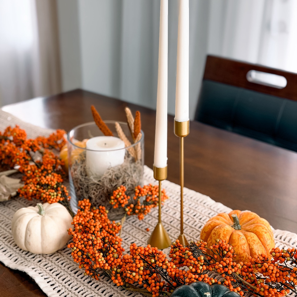 Fall centerpiece with gold taper candle holders, a berry garland and small pumpkins on a macrame runner