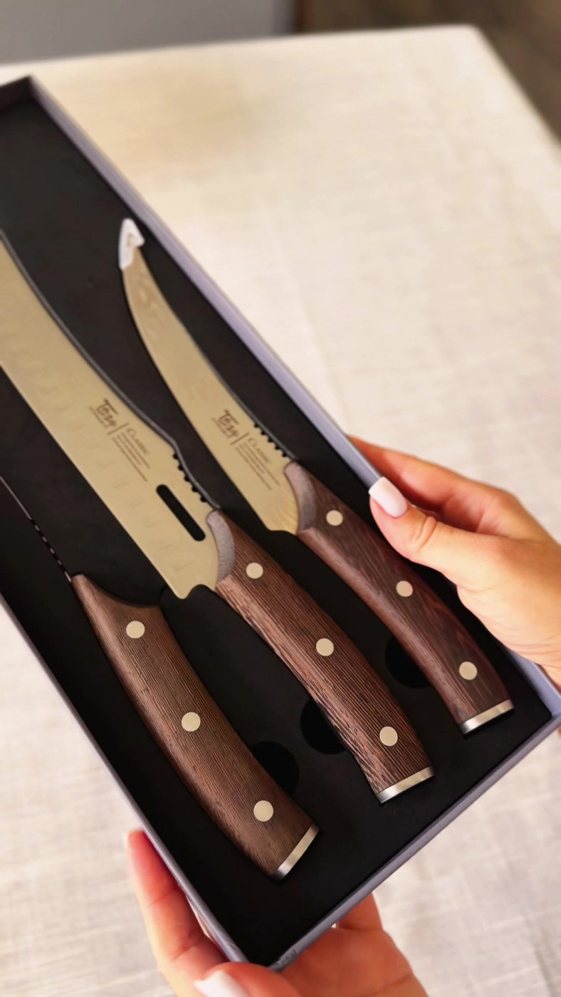A hand holding the open SYOKAMI butcher knife set showing three knives with dark wooden handles in a black presentation box